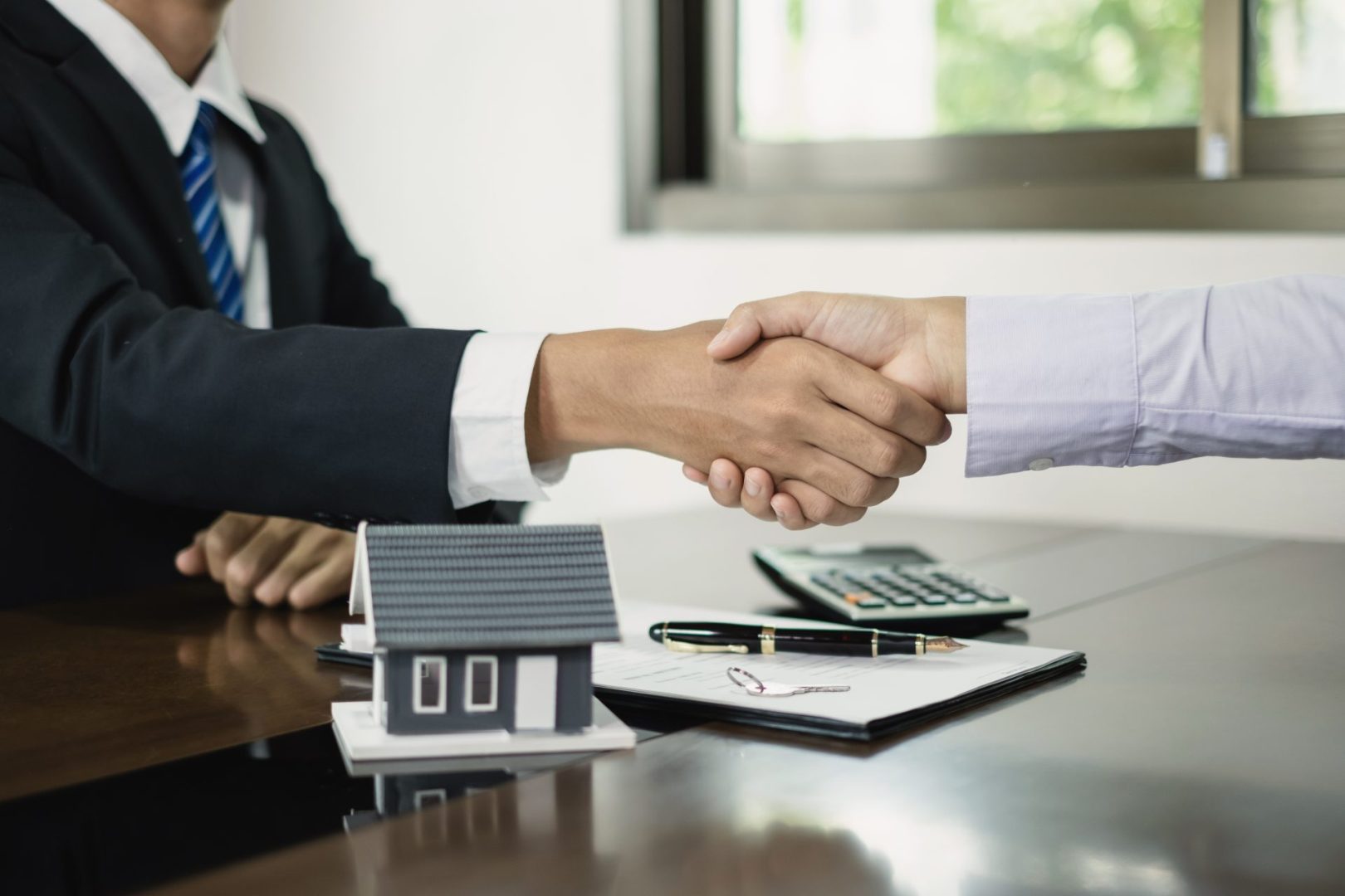 Businessmen and mortgage broker shake hands after completing negotiations to buy a home. Small decorative house, calculator and other stationery nearby.
