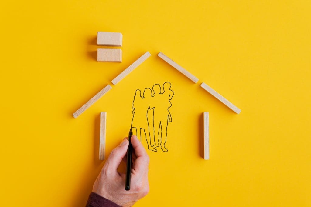 Male hand drawing silhouette of a family with black marker inside a house made of wooden blocks. Over yellow background.