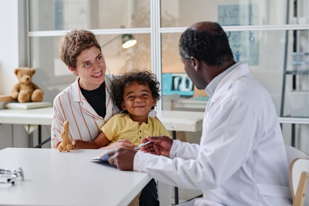Young woman visiting pediatrician with her little son, they are sitting at table and talking to a doctor