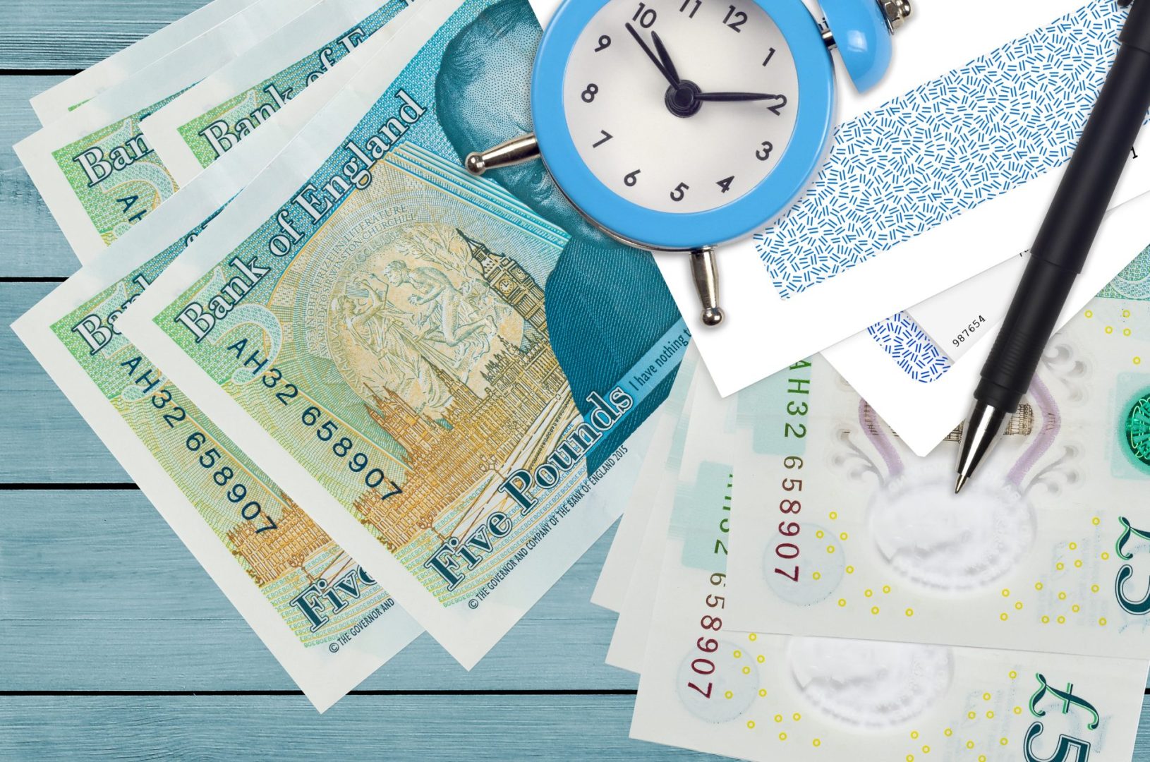five pound notes, envelopes, a blue clock and a pen sprawled out on a blue wooden surface