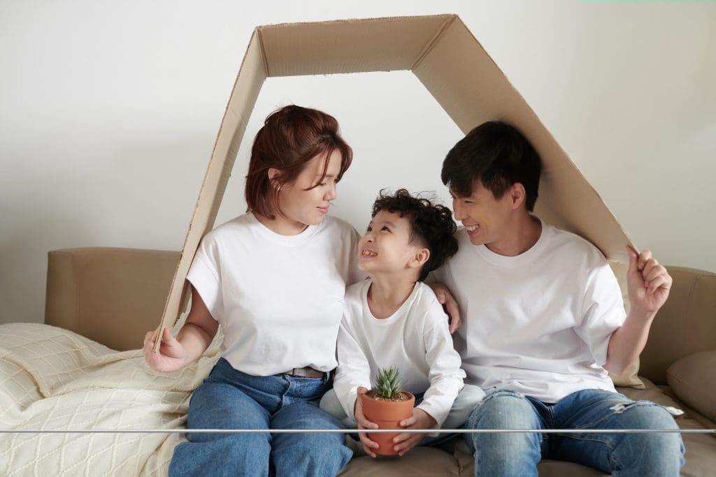 Cheerful little kid with flower pot looking at parent when they are sitting under cardboard roof symbolising house