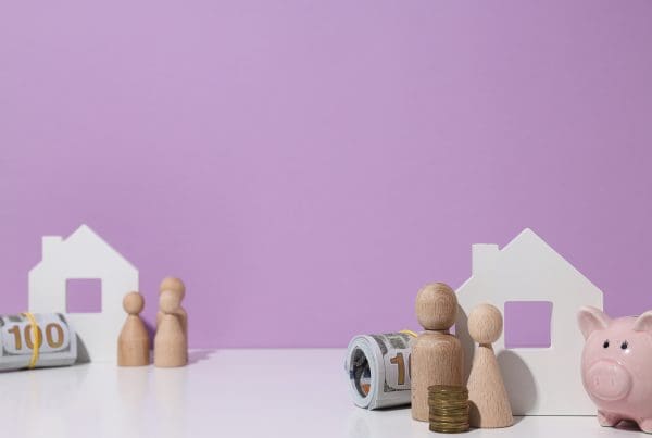 wooden houses, figures of people, piggy banks and rolls of cash on a pink/purple backgfround